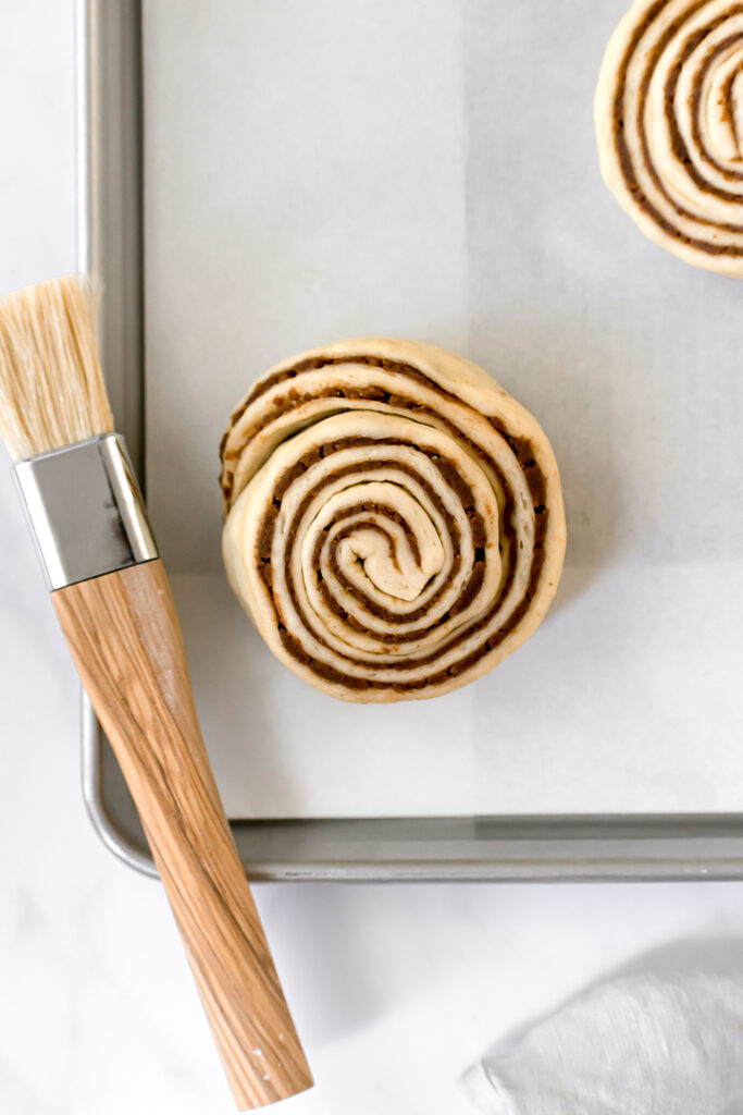 one cinnamon bun on baking sheet with pastry brush
