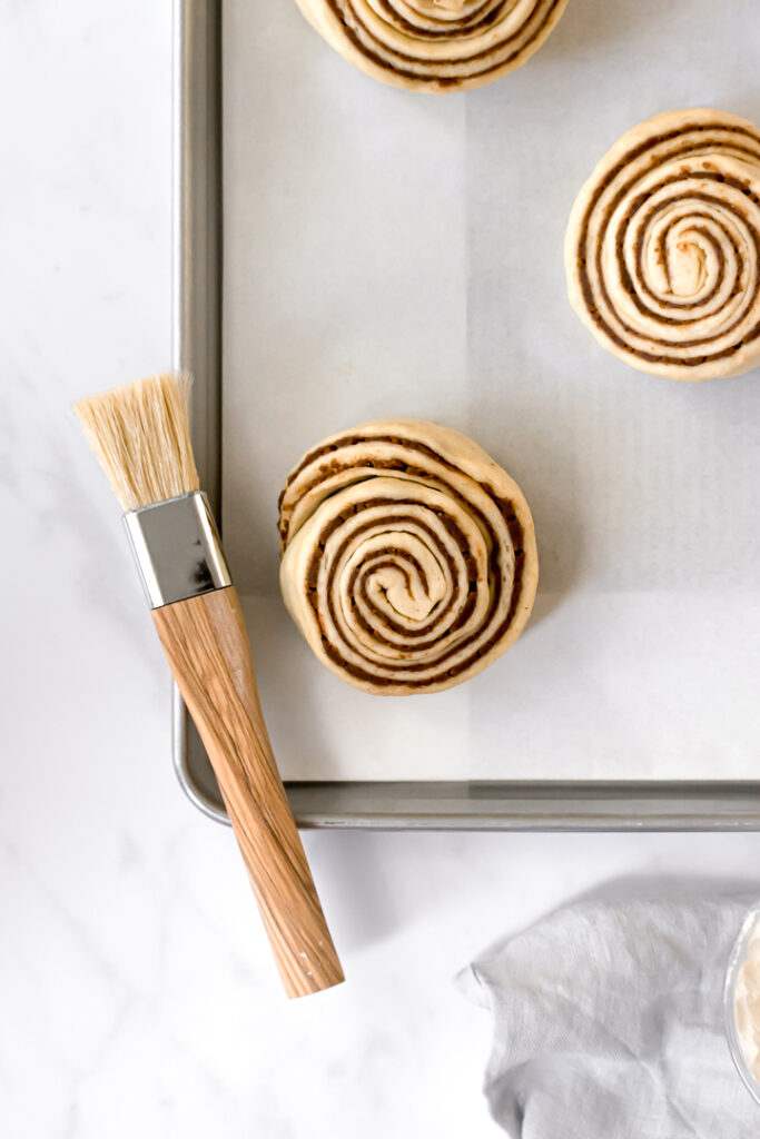 three cinnamon buns on baking sheet with pastry brush
