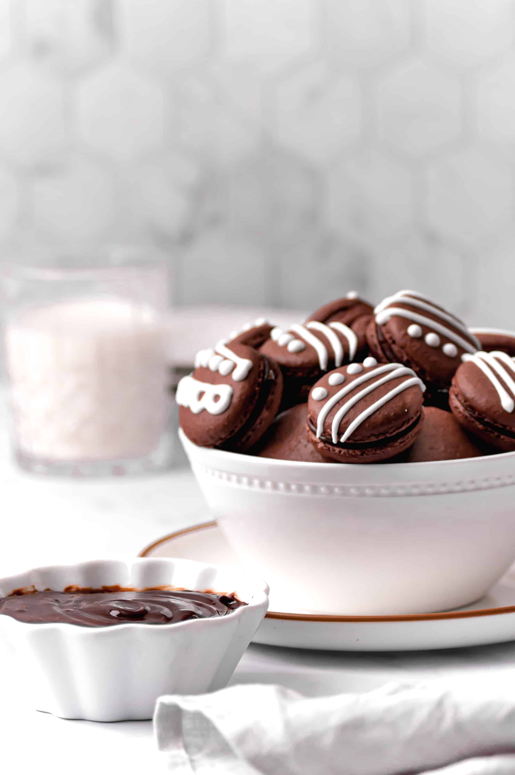 chocolate macarons decorated with royal icing in a bowl with glass of milk in background.