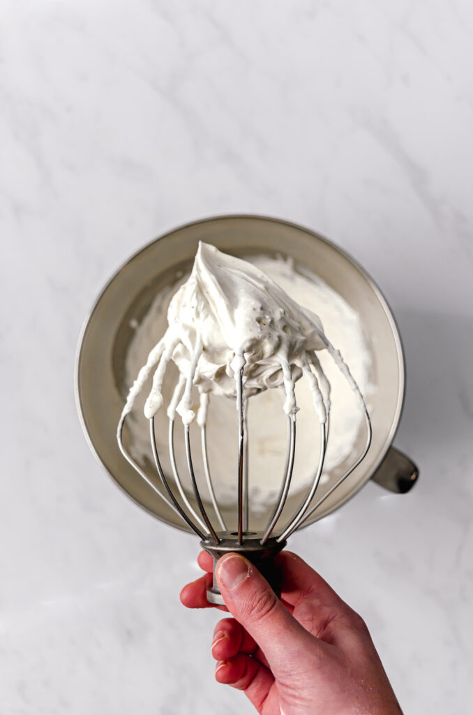 large whisk with mascarpone frosting held up above the bowl of frosting