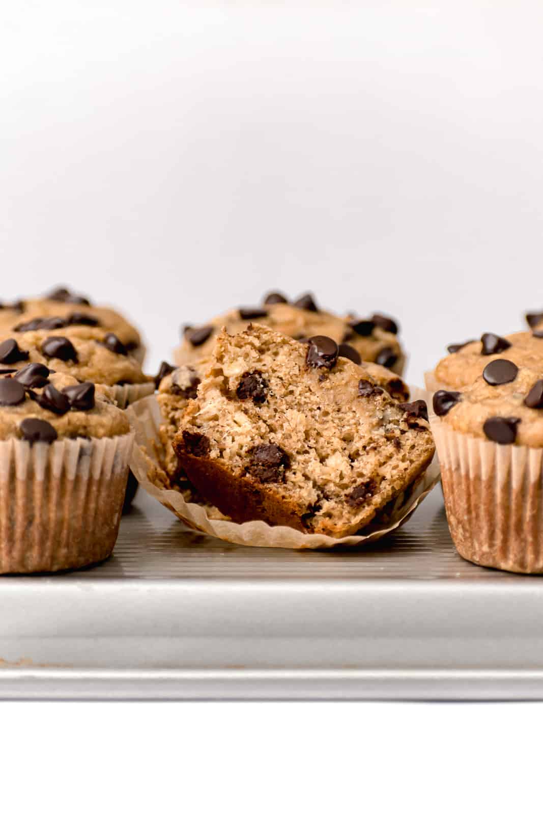 banana tahini chocolate chip muffins on silver baking sheet with one muffin cut in half.