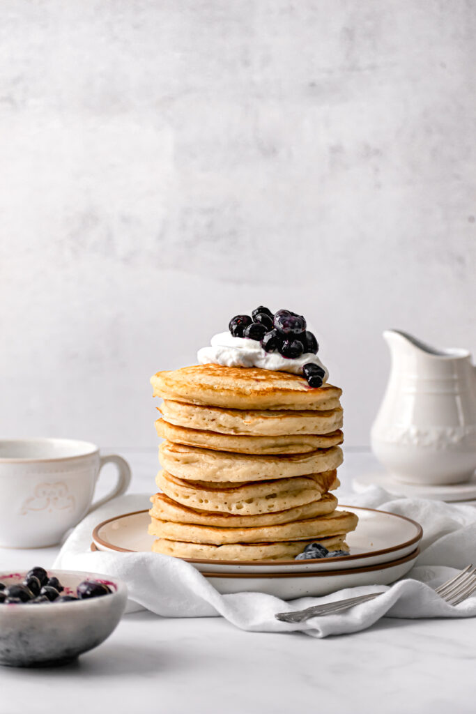 large stack of fluffy pancakes on white plate with whip cream and blueberry jam