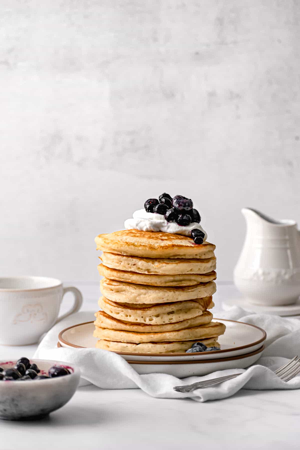 large stack of fluffy pancakes on white plate with whip cream and blueberry jam.