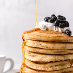 close up of stacked buttermilk pancakes with whip cream and blueberries and maple syrup drizzled on top