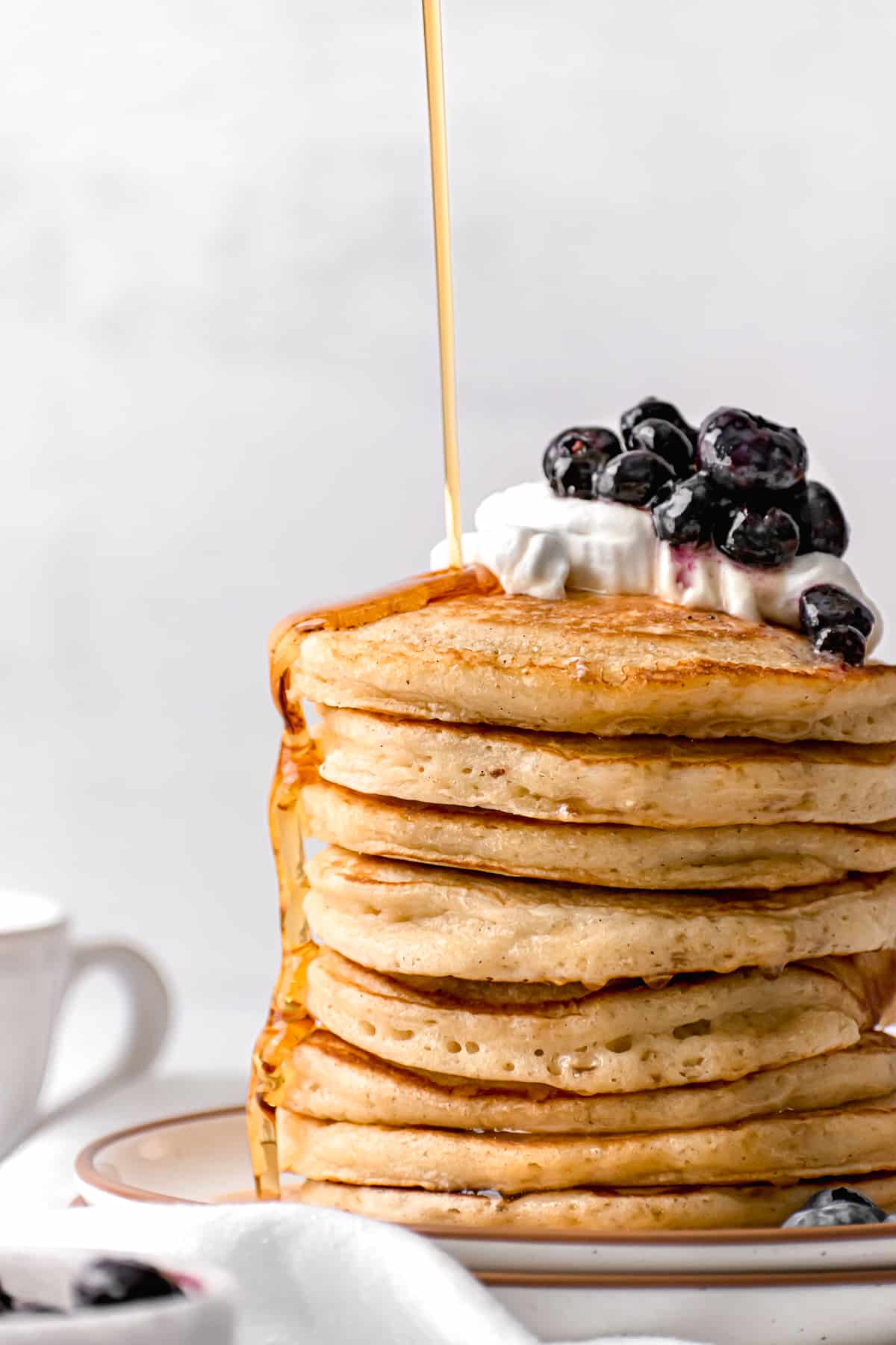 close up of stacked buttermilk pancakes with whip cream and blueberries and maple syrup drizzled on top.