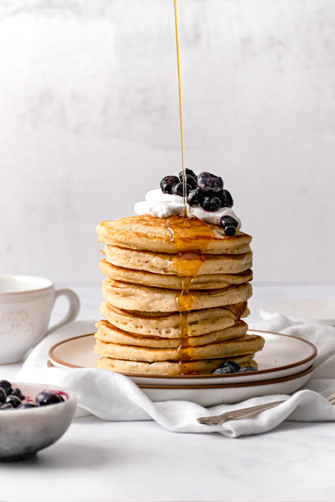 tall stack of fluffy buttermilk pancakes with maple syrup being drizzled on top