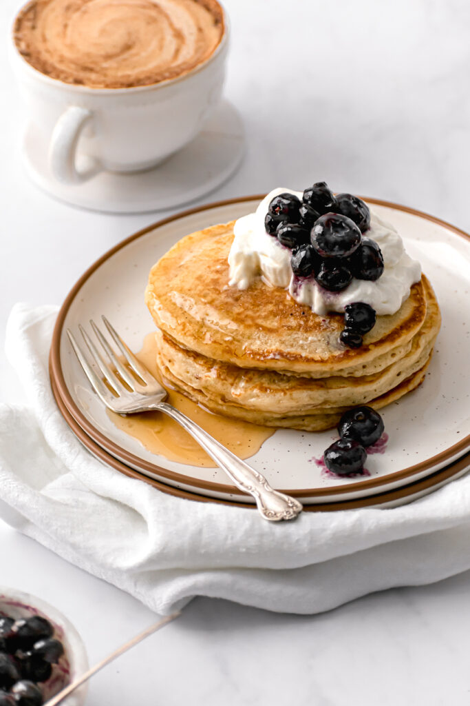 fluffy pancakes with whip cream, blueberries, and maple syrup on white plate with cup of coffee