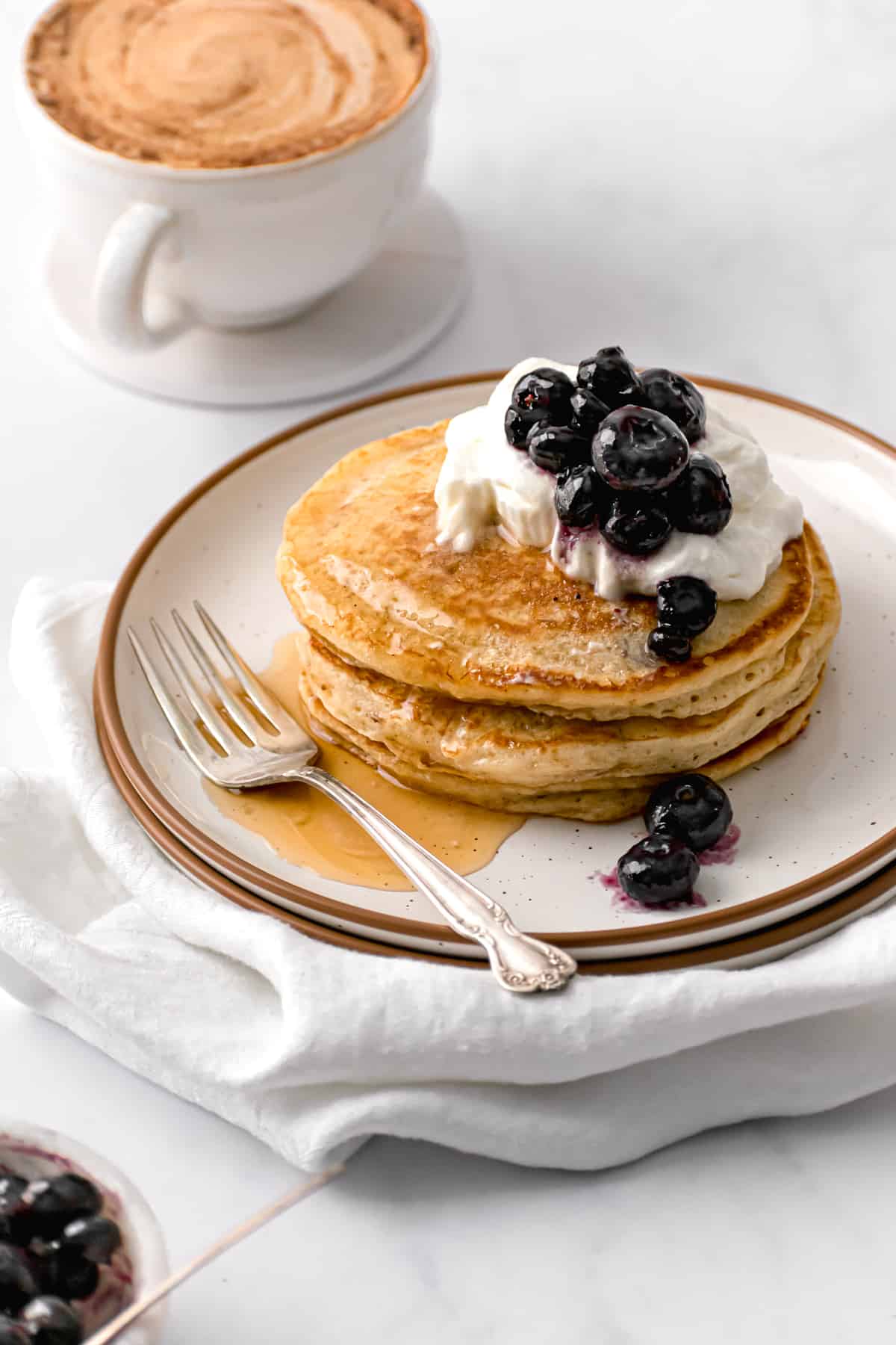 fluffy pancakes with whip cream, blueberries, and maple syrup on white plate with cup of coffee.