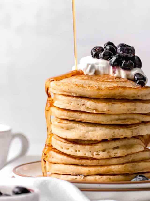 close up of stacked buttermilk pancakes with whip cream and blueberries and maple syrup drizzled on top