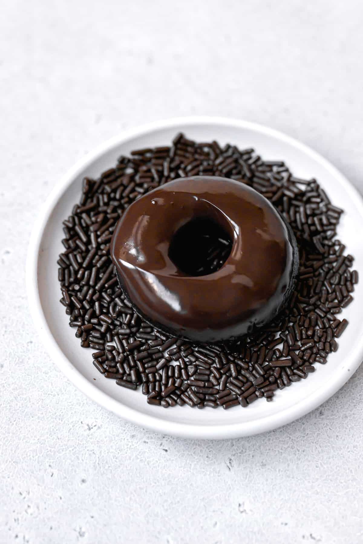 chocolate donut dipped in glaze and sitting on a white plate full of sprinkles.