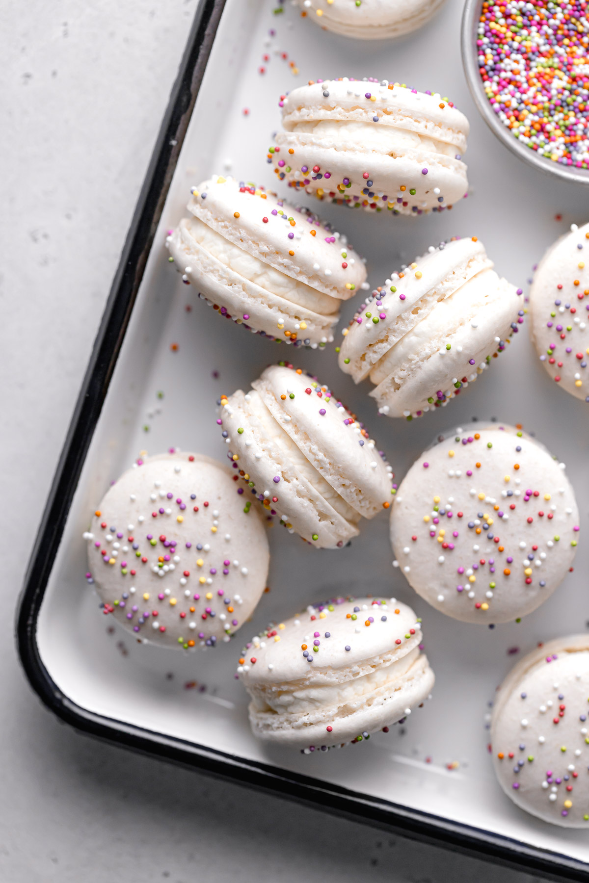 birthday cake macarons with rainbow sprinkles and whipped cream cheese frosting arranged in white tray.