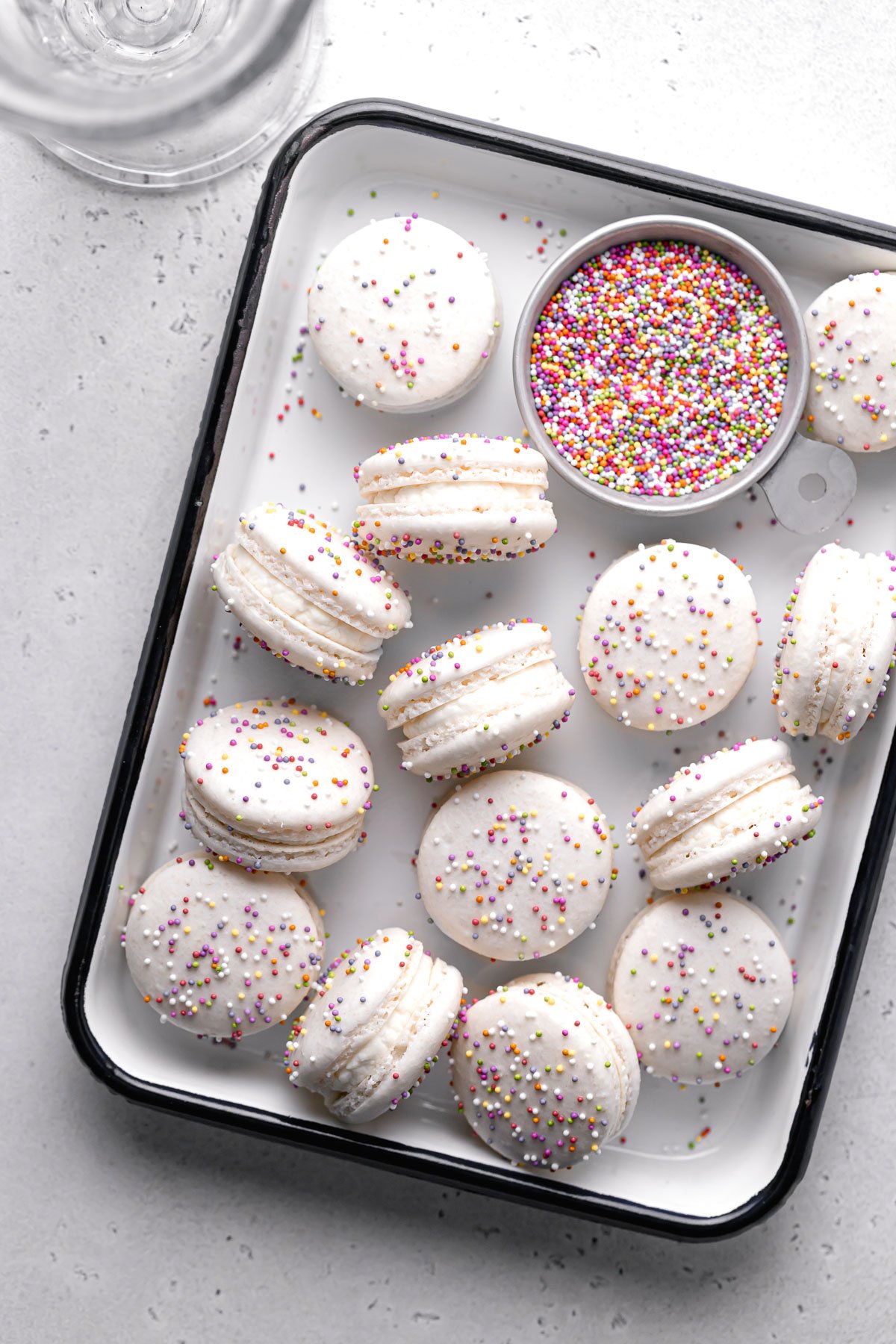 birthday cake macarons with a bowl of rainbow sprinkles in white tray.