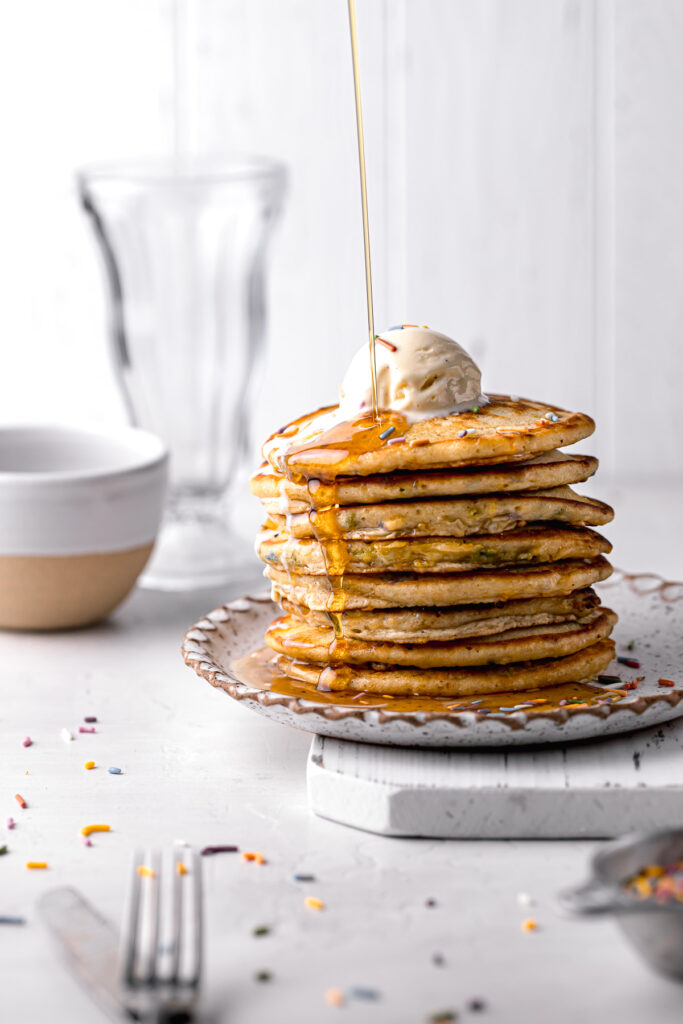 funfetti pancakes topped with ice cream and maple syrup being poured on top