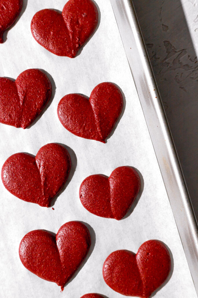 red macaron batter piped into heart shapes on baking sheet