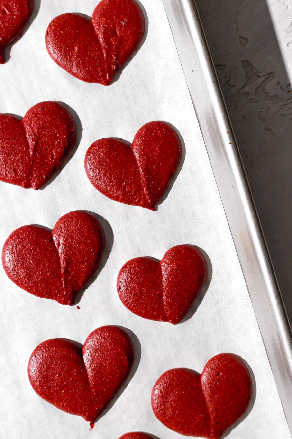 red macaron batter piped into heart shapes on baking sheet.