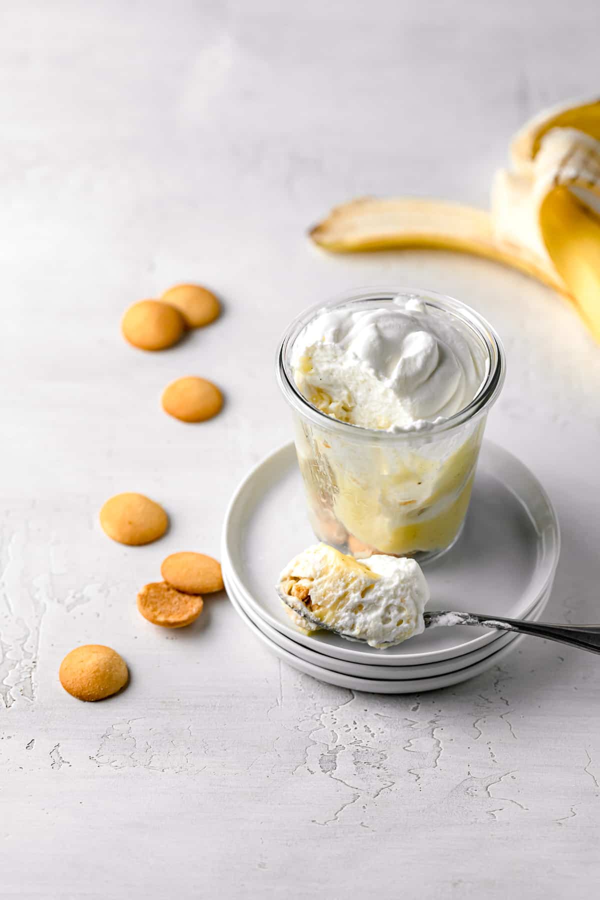 banana pudding jar on stacked plates with spoon, nilla wafers spread around, and open banana. 