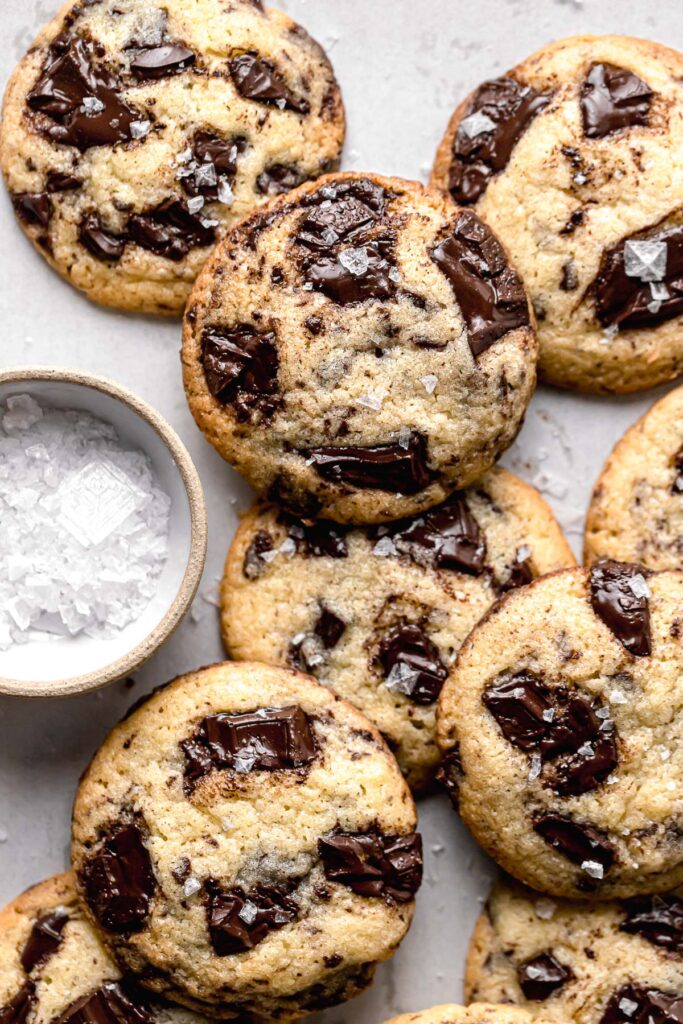 chocolate chip cookies without brown sugar piled on parchment lined baking sheet with bowl of sea salt