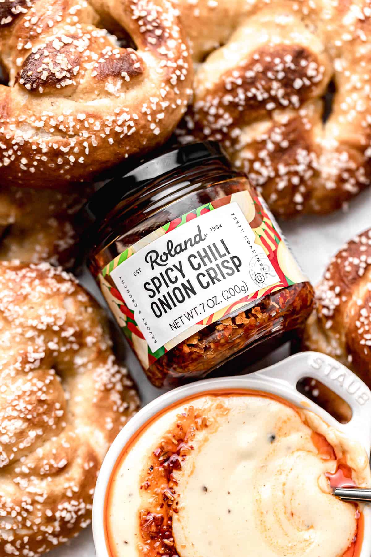 close up of a jar of Roland spicy chili onion crisp surrounded by baked soft pretzels and cheese dip.