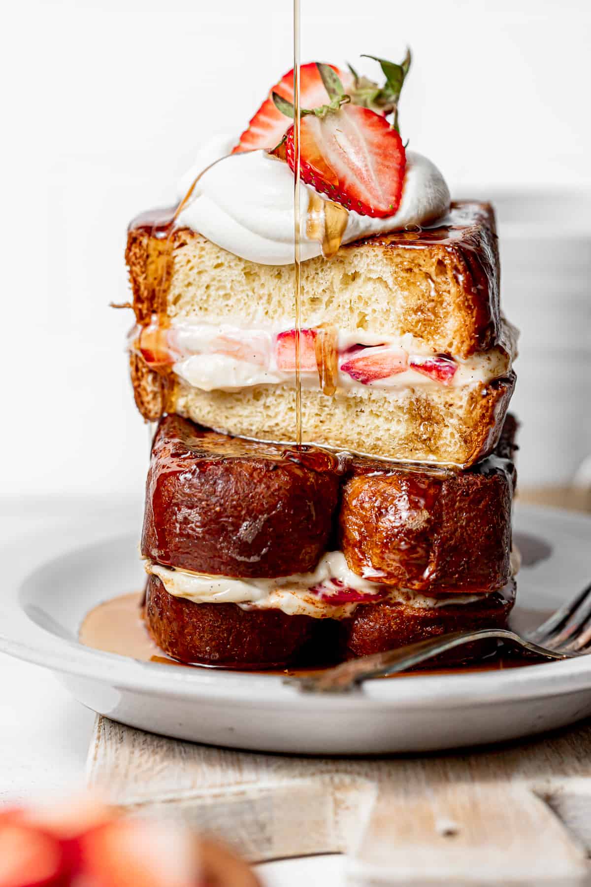 strawberries and cream stuffed French toast on white plate with maple syrup being poured on top.
