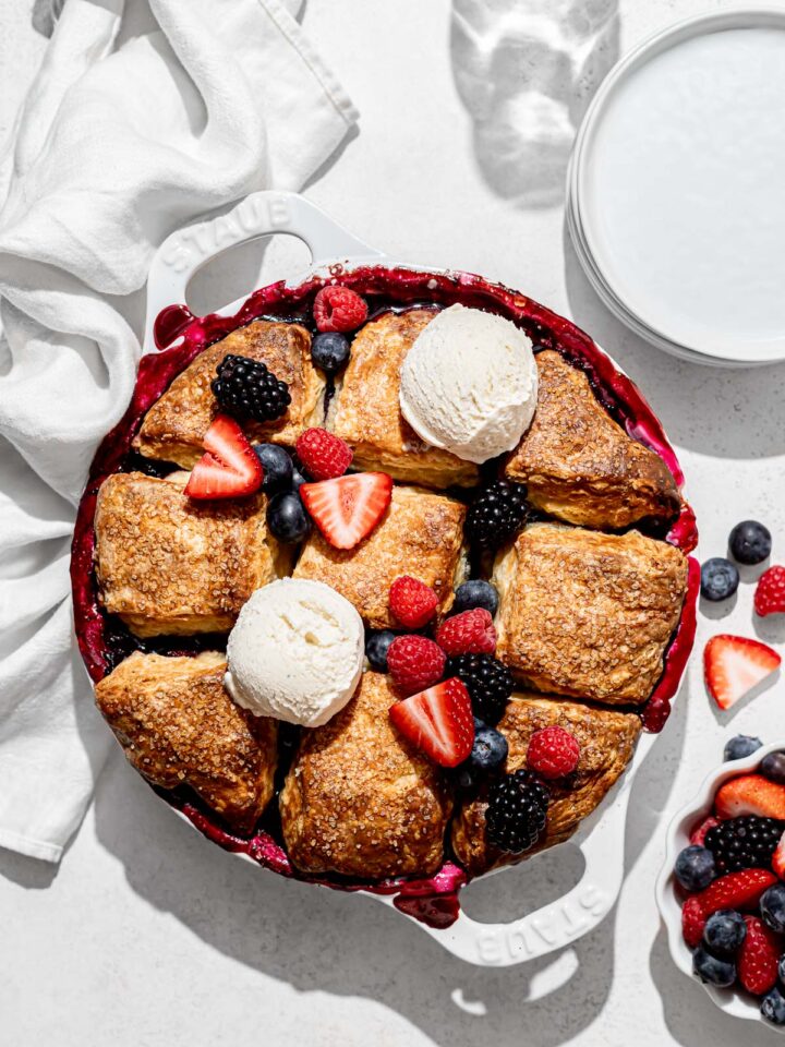 mixed berry cobbler with biscuits, fresh fruit, and ice cream on top.
