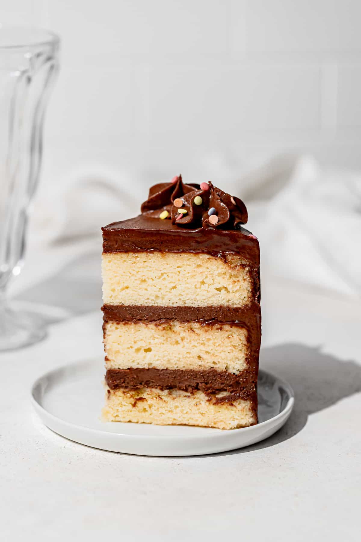 slice of yellow cake with chocolate frosting standing up on white plate. 