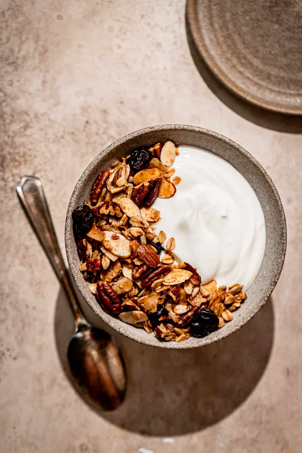 maple nut granola in a bowl of yogurt on a beige background with a spoon resting by the bowl.