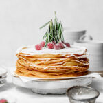 maple eggnog crepe cake topped with powdered sugar on white wooden cake stand.