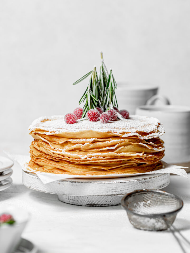 maple eggnog crepe cake topped with powdered sugar on white wooden cake stand.