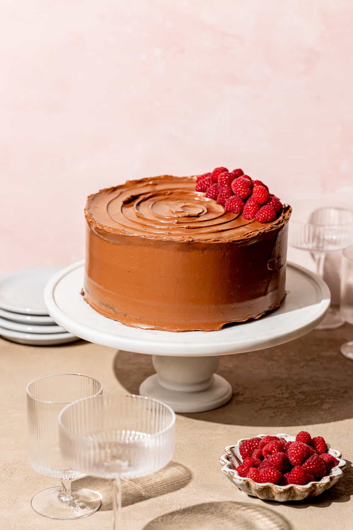 frosted and decorated chocolate raspberry cake on white cake stand with a pink backdrop.
