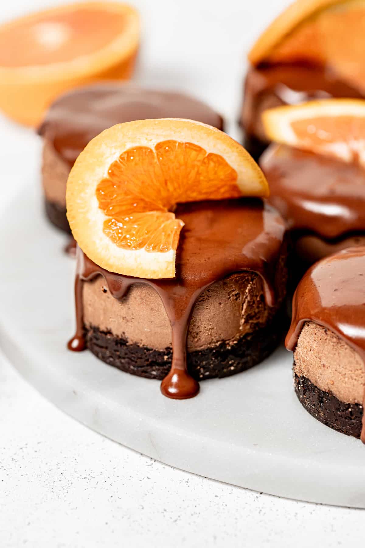 close up of mini chocolate orange cheesecakes with orange slices on marble slab.