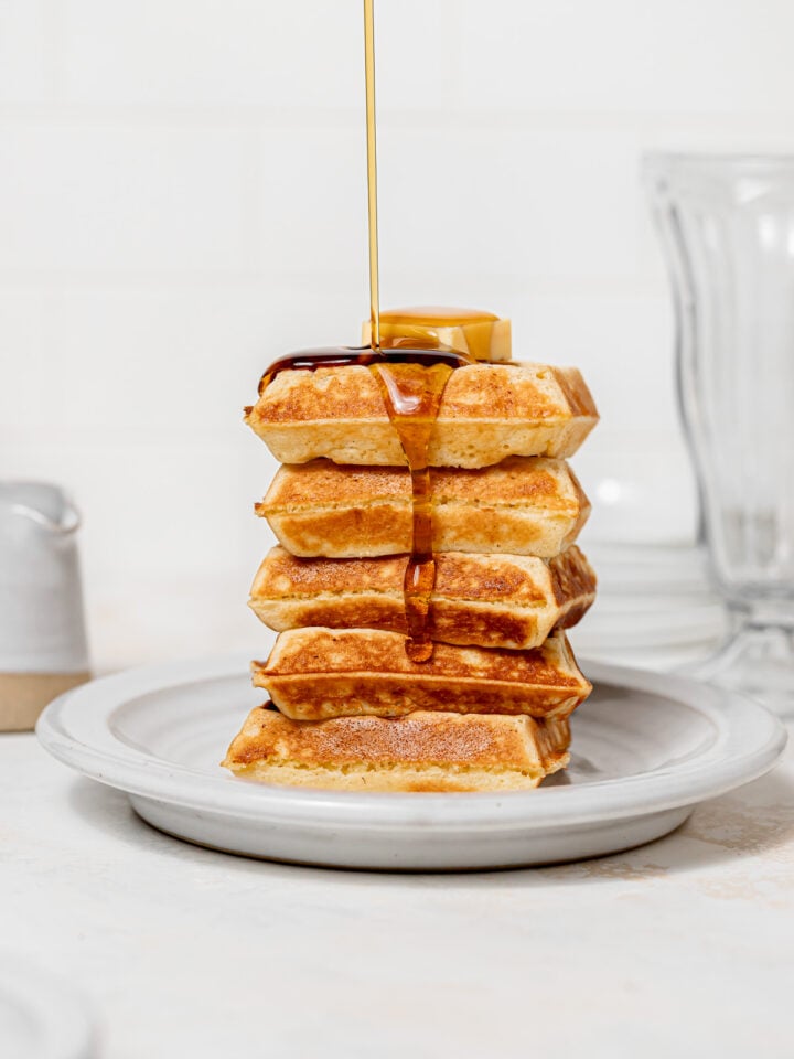 yeasted belgian waffles stacked with maple syrup being poured on top.